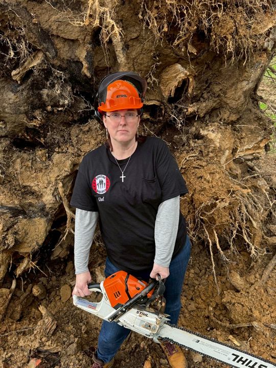 A volunteer in a hard hat holds a chainsaw in front of an uprooted tree