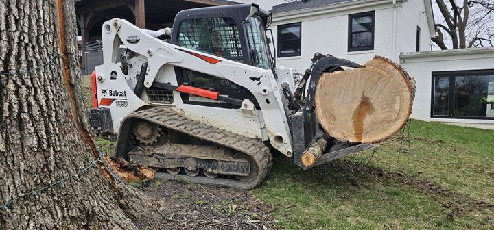 A tracked loader carries a large log section past a house