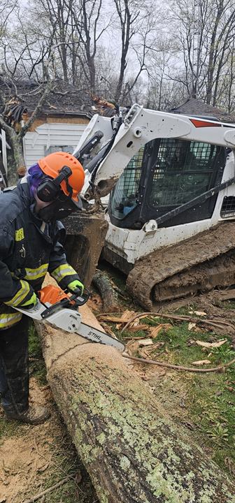 A volunteer cuts a fallen tree beside a skid steer