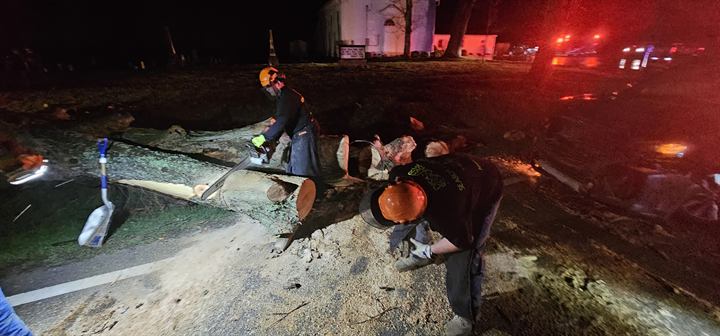 Volunteers clear a downed tree at night by the light of emergency vehicles