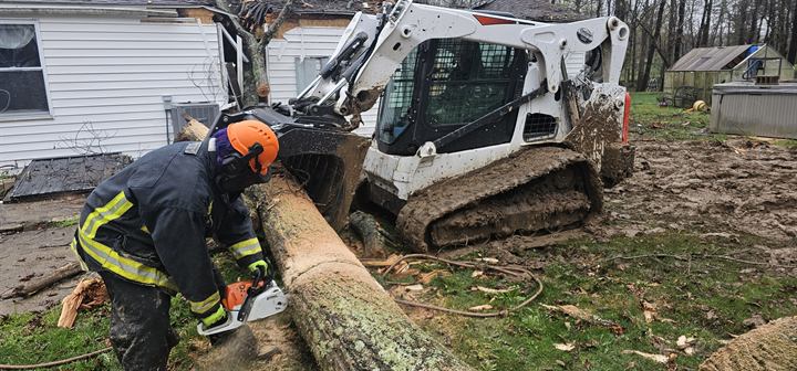 A volunteer in turnout gear cuts a fallen tree beside a skid steer