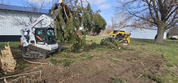 A tracked loader clears a downed tree beside a farm building